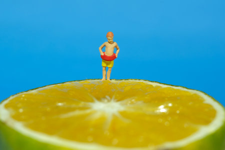 Miniature people toy figure photography. Young boy, kids, with rubber ring tube standing at the edge of slice orange fruit. Isolated on blue background. Image photoの写真素材
