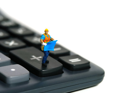 Miniature people toy figure photography. Project budget cost calculation concept. A construction worker with blueprint building standing above calculator. Isolated on white background. Image photoの写真素材