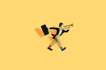 Miniature people toy figure photography. A clown wearing black suit and hat blow the trumpet on yellow background. Image photoの写真素材