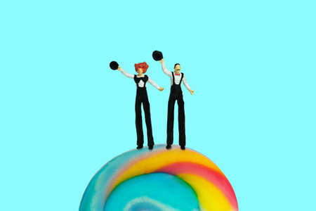 Miniature people toy figure photography. Front view, full body of a tall men clown circus standing above rainbow candy while waving hand and holding black hat. Isolate on blue background. Image photoの写真素材
