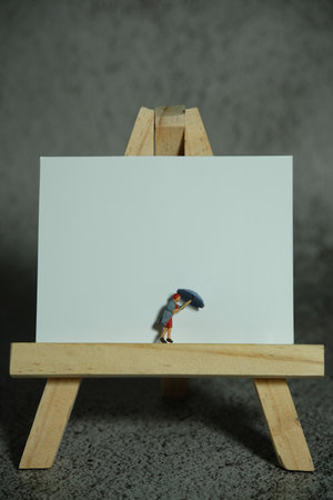 Miniature people toy figure photography. Women standing above wooden stand on blank desk calendar. Grey cloudy background. Image photoの写真素材