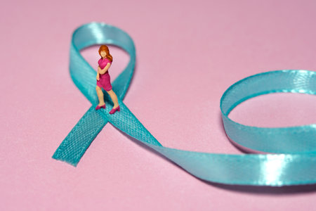Miniature people figure toys photography. Ovarian and breast cancer awareness day concept. Girl, teenage, woman standing above green turquoise teal ribbon. Image photoの写真素材