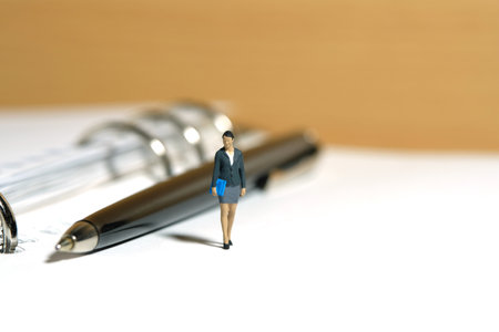 Miniature people toy figure photography. Minutes of meeting, schedule, task checklist concept. A businesswoman standing walking above notebook with pen. Isolated on white background. Image photoの写真素材