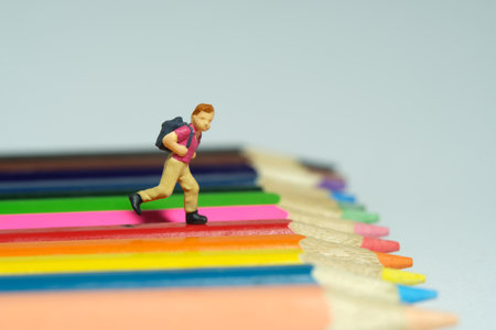 Miniature tiny people toys photography. A boy with school backpack running above colorful pencil. Isolated on grey background. Image photoの写真素材