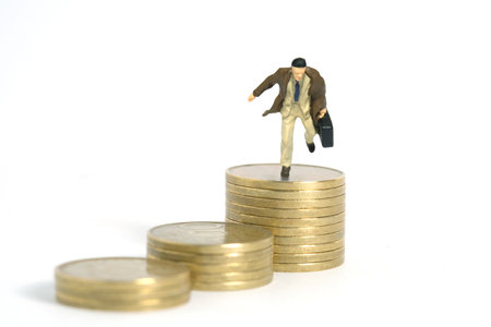 Miniature tiny people toy figure photography. A businessman wearing coat jumping above golden coin stack. Isolated on white background. Image photoの写真素材