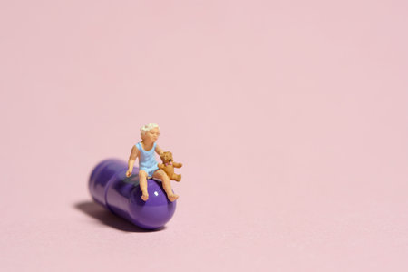 Miniature tiny people toy figure photography. A girl toddler sitting above purple medicine pill with teddy bear. Isolated on pink background. Image photoの写真素材