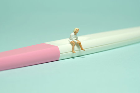 Miniature tiny people toy figure photography. A girl teenager sitting above pregnancy test pack tool. Isolated on a green background. Image photoの写真素材