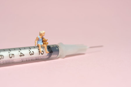 Miniature tiny people toy figure photography. A girl toddler sitting above needle syringe holding teddy bear doll. Isolated on pink background. Image photoの写真素材