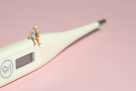 Miniature tiny people toy figure photography. A girl toddler sitting above white thermometer holding...