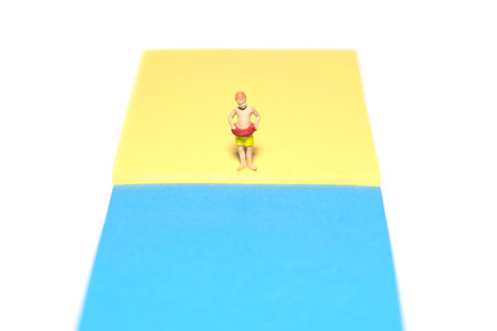 Creative miniature people toy figure photography. Sticky notes installation. Young boy, kids, with rubber ring tube standing at beach shore. Isolated on white background. Image photoの写真素材