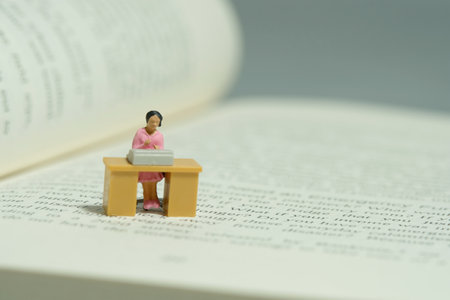 Miniature tiny people toy figure photography. Literature research. A woman seat on desk above opened book. Image photoの写真素材