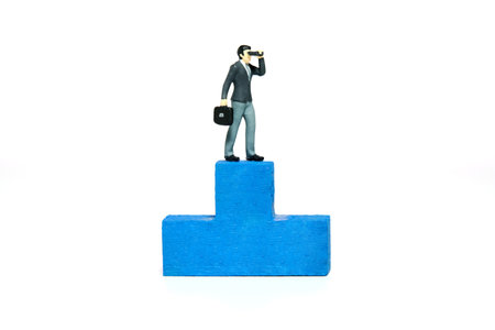 Miniature tiny people toy figure photography. Winning tactics concept illustration. A businessman using binocular telescope standing above blue podium. Isolated on a white background. Image photoの写真素材