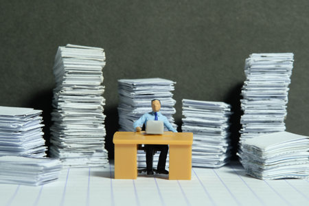 Miniature tiny people toy figure photography. Overload and work overtime concept. A businessman seat on a desk with pile of document. Image photoの写真素材