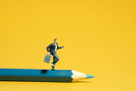 Miniature tiny people toys photography. A boy student running above blue pencil. Isolated on yellow background. Image photoの写真素材