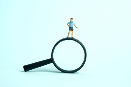 Miniature people toy figure photography. Children curiosity concept. A boy standing in front of black magnifier glass. Isolated on blue background. Image photoの写真素材