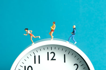 Miniature people toy figure photography. Running daily routine concept. A team group runner jogging above clock, isolated on blue background. Image photoの写真素材