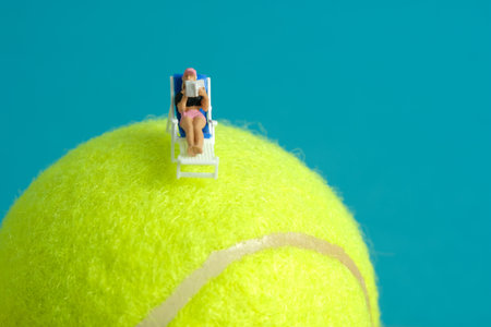 Miniature people toy figure photography. Girl relaxing seat at beach chair while reading book above tennis ball. Image photoの写真素材