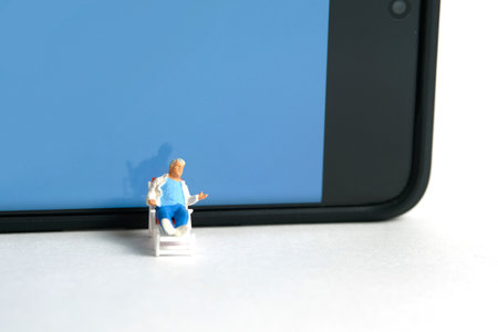 Miniature people toy figure photography. Virtual travel concept, Grandfather relaxing seat while reading book at chair with smartphone, isolated on white background. Image photoの写真素材