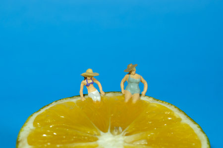 Miniature people toy figure photography. Two women relaxing above orange fruit. Isolated on blue background. Image photoの写真素材