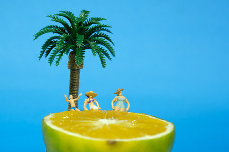 Miniature people toy figure photography. Two women and a boy relaxing above slice orange fruit under coconut tree. Isolated on blue background. Image photoの写真素材