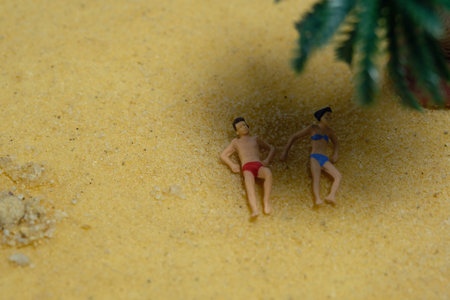 Miniature people toy figure photography. Top eagle view of men and girl couple relaxing, lying at beach under a coconut tree. Image photoの写真素材