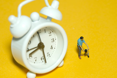 Miniature people toy figure photography. Time for children concept. A father and son standing in front of white clock. Isolated yellow background. Image photoの写真素材