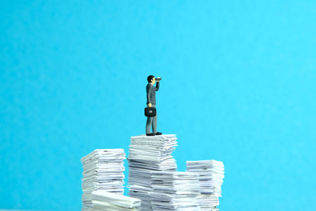 Miniature tiny people toy figure photography. Project task observation concept illustration. Businessmen using binocular standing above file pile. Isolated on a blue background. Image photoの写真素材
