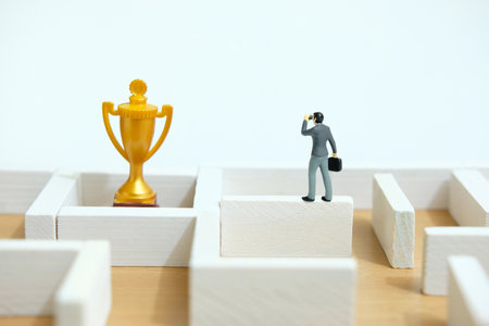 Miniature tiny people toy figure photography. Business barrier concept illustration. A businessman using binocular telescope standing above maze labyrinth wall looking golden trophy. Image photoの写真素材