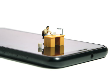 Miniature tiny people toy figure photography. Work from anywhere, mobile worker concept. A businessmen seat on his desk, above smartphone. Isolated on a white background. Image photoの写真素材