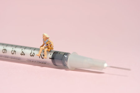 Miniature tiny people toy figure photography. A girl toddler sitting above needle syringe holding teddy bear doll. Isolated on pink background. Image photoの写真素材