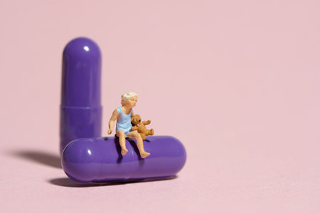 Miniature tiny people toy figure photography. A girl toddler sitting above purple medicine pill with teddy bear. Isolated on pink background.の写真素材