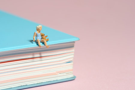 Miniature tiny people toy figure photography. A girl toddler sitting above book holding teddy bear doll. Isolated on pink background. Image photoの写真素材