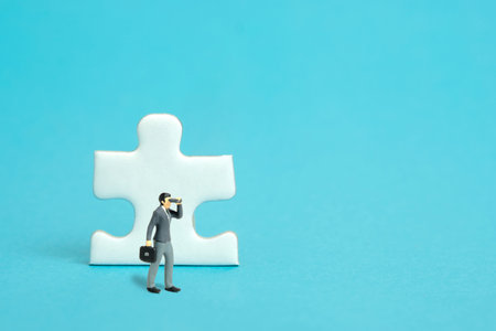 Miniature tiny people toy figure photography. Businessmen standing above jigsaw puzzle piece using binoculars. isolated on blue background. Image photoの写真素材