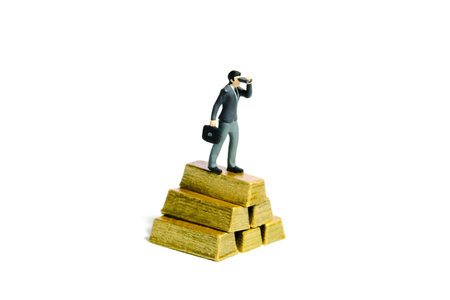Miniature tiny people toy figure photography. Investment concept illustration. Businessman using binocular telescope standing above gold bar stack. Isolated on a white background. Image photoの写真素材