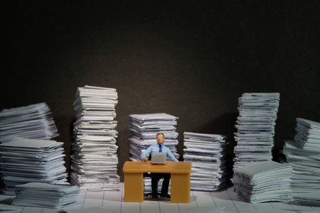 Miniature tiny people toy figure photography. Overload and work overtime concept. A businessman seat on a desk with pile of document, working until night at dark room with spotlight. Image photoの写真素材