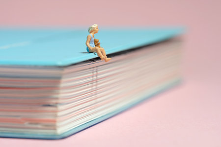 Miniature tiny people toy figure photography. A girl toddler sitting above book holding teddy bear doll. Isolated on pink background. Image photoの写真素材