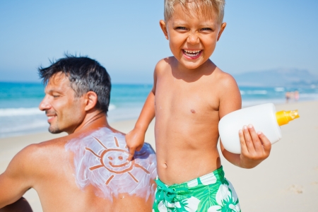 Adorable boy at tropical beach applying sunblock cream on a father s back の写真素材