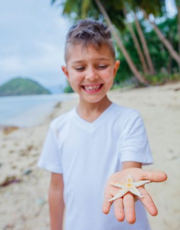 Boy holding a starfish on tropical sandy beach. Focus on the starfishの写真素材