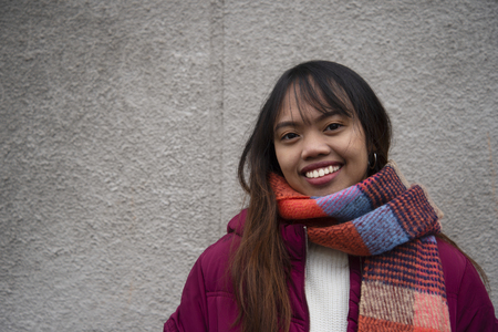 20s smile Filipino young teen woman Asian portrait with red coat and scarf with grey wall backgroundの写真素材