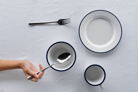 set of white dish bowl cup on food dining table with fork and woman hand holding metal spoon on tablecloth backgroundの写真素材
