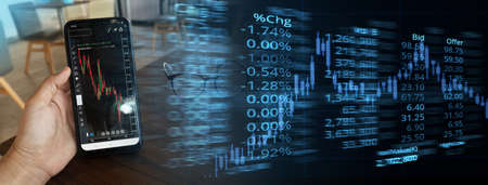 hand holding smart phone with red green graph of stock market display and ticker board finance and...