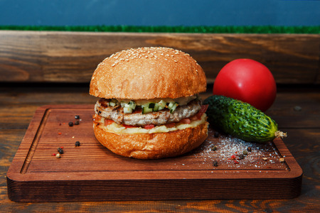 One burger on a wooden board. Burger with pork cutlet. Next to him lies a cucumber and a tomato. Sprinkled spicesの写真素材
