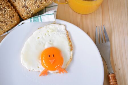 A fried egg lies on a plate, next to it are bread rolls and cutlery. The egg looks like a chick, with two grains of eyes and pieces of carrot as mouth and feetの写真素材