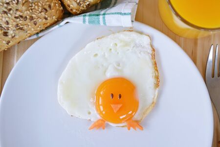 A fried egg lies on a plate, next to it are bread rolls and cutlery. The egg looks like a chick, with two grains of eyes and pieces of carrot as mouth and feetの写真素材