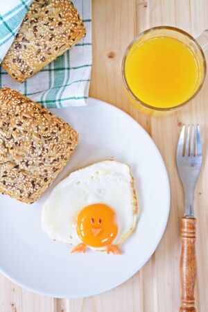 A fried egg lies on a plate, next to it are bread rolls and cutlery. The egg looks like a chick, with two grains of eyes and pieces of carrot as mouth and feetの写真素材