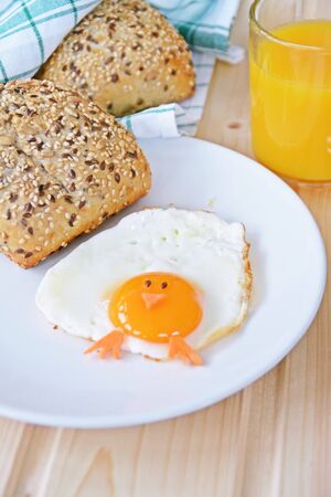 A fried egg lies on a plate, next to it are bread rolls and cutlery. The egg looks like a chick, with two grains of eyes and pieces of carrot as mouth and feetの写真素材