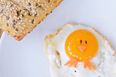 A fried egg lies on a plate, next to it are bread rolls and cutlery. The egg looks like a chick, with two grains of eyes and pieces of carrot as mouth and feetの写真素材