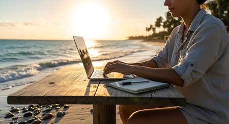 Young woman focused on laptop, working remotely from a beautiful tropical beach at sunset.の素材