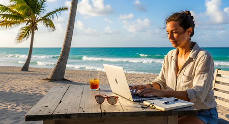 Young woman working on laptop at a beach with palm trees and ocean waves.の素材