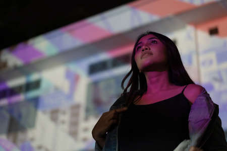 Young hipster woman standing outdoors at night in front of light projector with pink light projected on her body and faceの写真素材